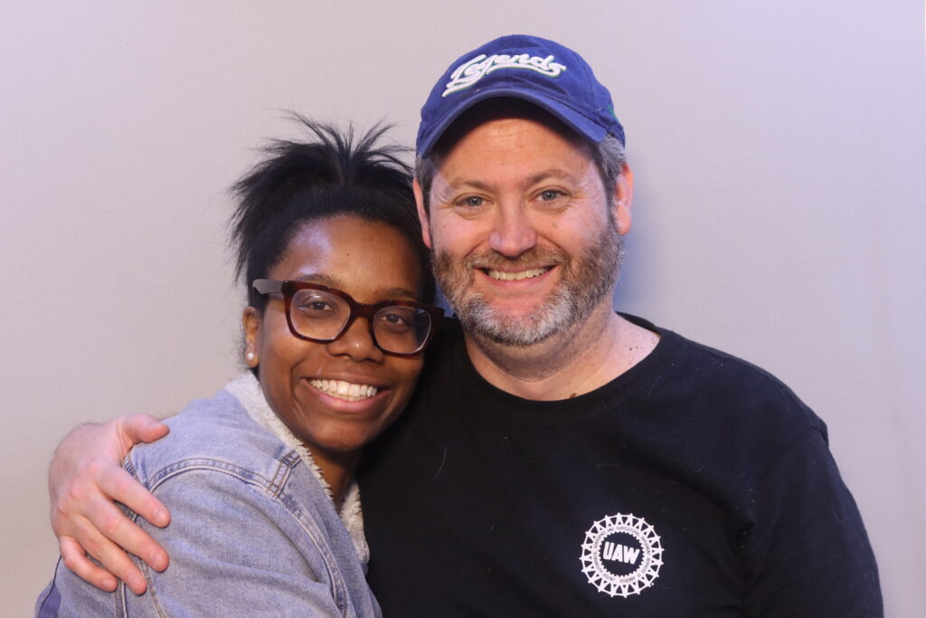A Black woman and a white man with a beard smile at the viewer, their arms around each other's shoulders.